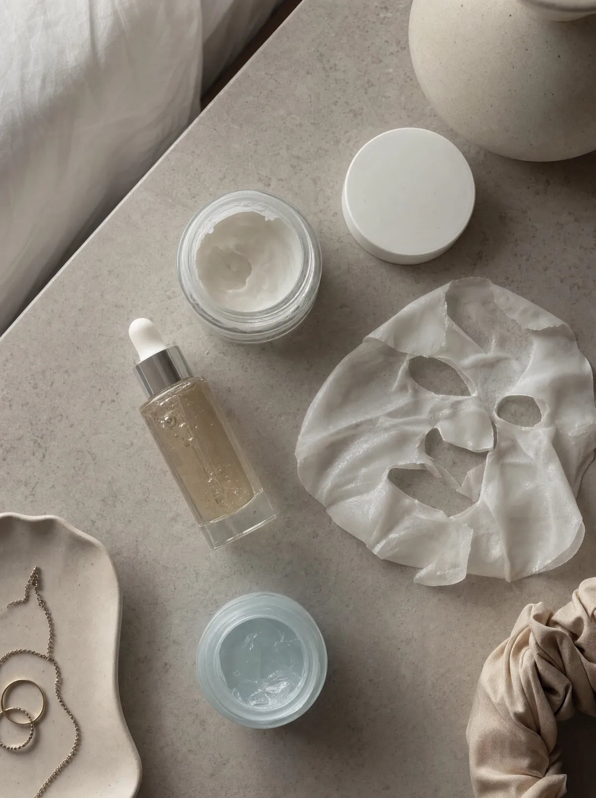 A half-used night cream, a serum dropper, a peeled sheet mask and a jelly jar scattered on a nightstand